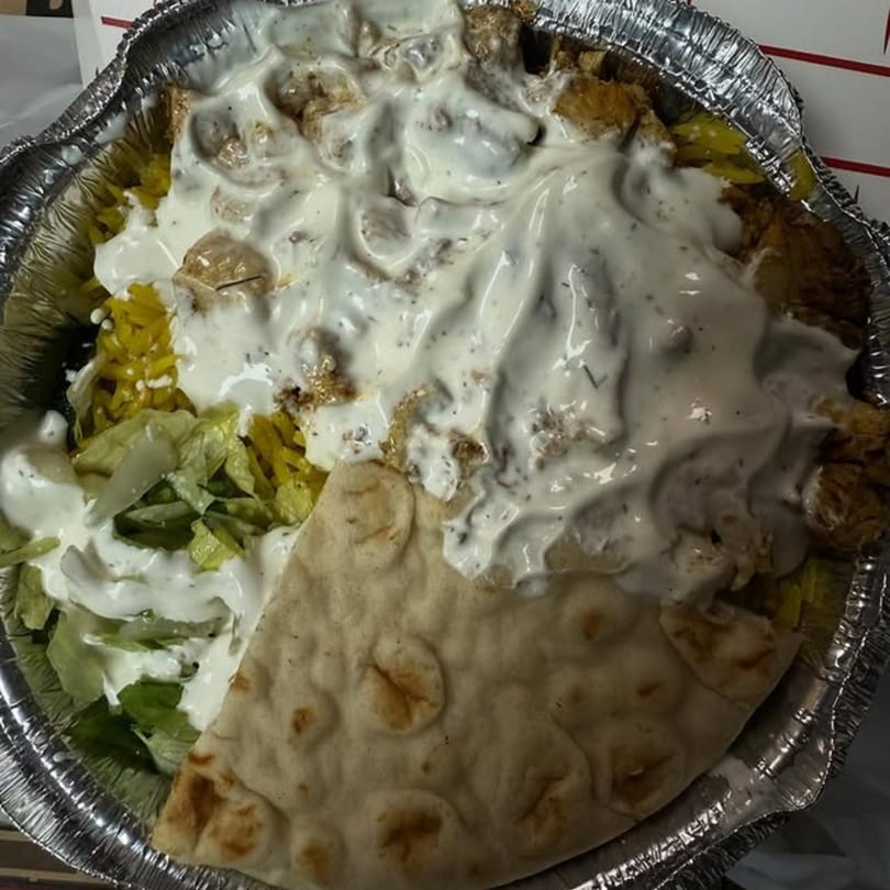 A foil container holds a gyro platter with meat, rice, lettuce, white sauce, and pita bread.