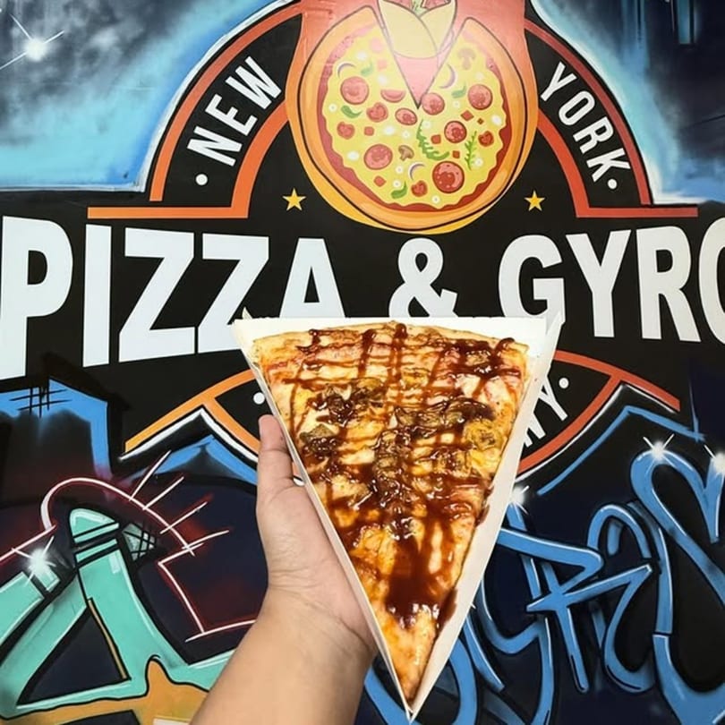 Hand holding a large slice of BBQ chicken pizza with a graffiti-style NY Pizza & Gyro sign in the background.